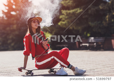 Vaping girl. Young woman sitting on skateboard and vape e-cig. Pretty young female in black hat, red 51699797