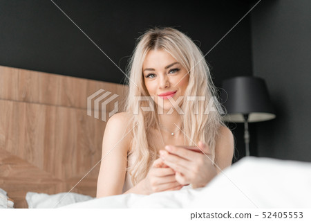 Use the phone. Checks email and social media messages. Young woman wake up, sits on comfortable bed Use the phone. Checks email and social media messages. Young woman wake up, sits on comfortable bed 52405553