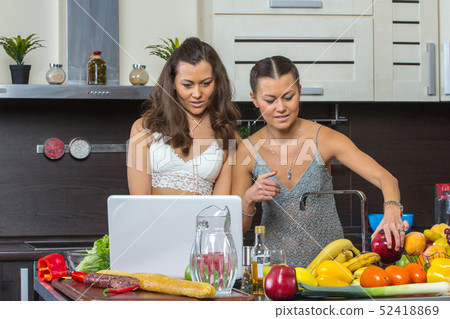 Two attractive women looking at laptop, standing in kitchen.  52418869