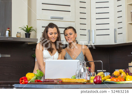 Two attractive twins women looking at laptop, standing in kitchen early morning. Two attractive twins women looking at laptop, standing in kitchen early morning. 52463216