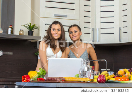 Two attractive twins women looking at laptop, standing in kitchen early morning. 52463285