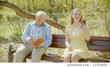 Old man is reading book in the park and young lady chatterbox interrupting him with loud chatting on 52554430