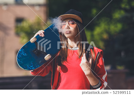 Vaping girl. Young woman with skateboard vape e-cig. Pretty young female in black hat, red clothing 52599511