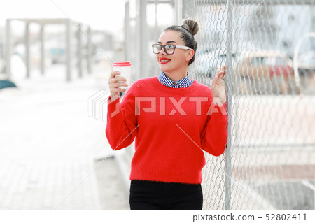 Beautiful woman in glasses, red sweater and red lipstick walking down the street with a glass of 52802411