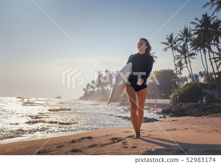 Beautiful young woman in a bathing suit walking along the ocean shore with a surfboard in her hands 52983174