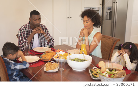 African American family with hands clasped and eyes closed praying together at dining table 53000171