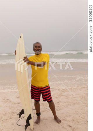 Happy senior male surfer standing with surfboard and looking at camera on the beach  53000192
