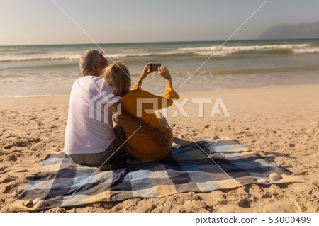 Senior couple taking selfie with mobile phone relaxing on picnic blanket at beach Senior couple taking selfie with mobile phone relaxing on picnic blanket at beach 53000499