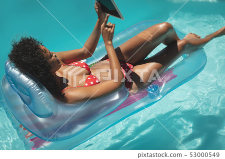 Young African American woman relaxing on pool lounger and using digital tablet 53000549