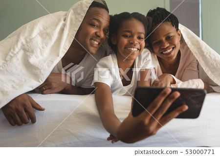 Happy African American family under blanket and taking selfie with mobile phone on bed Happy African American family under blanket and taking selfie with mobile phone on bed 53000771