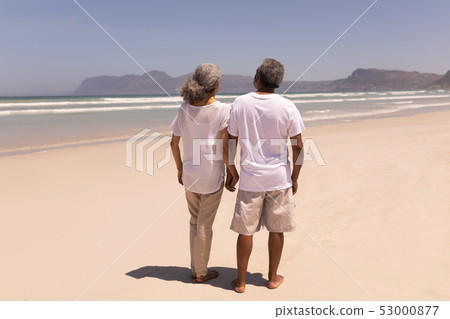 Senior couple holding hands and walking on beach with mountains in the background Senior couple holding hands and walking on beach with mountains in the background 53000877