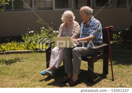 Front view of senior couple sitting on bench and using laptop in garden 53001287