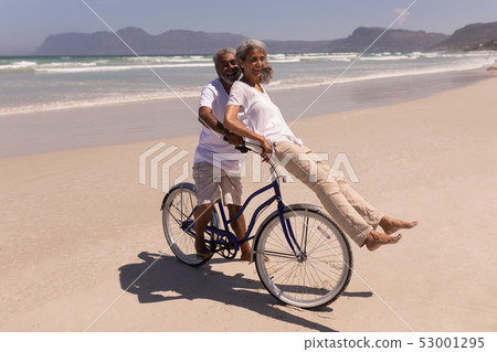 Senior man rides a bicycle with senior woman sitting on handlebar at beach Senior man rides a bicycle with senior woman sitting on handlebar at beach 53001295