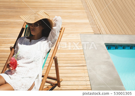 African American woman with cocktail glass relaxing on sun lounger in backyard 53001305
