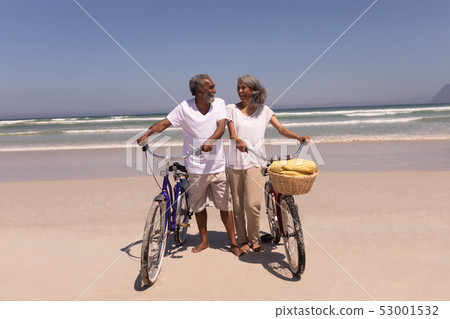 Senior couple walking with bicycle and looking each other on beach Senior couple walking with bicycle and looking each other on beach 53001532