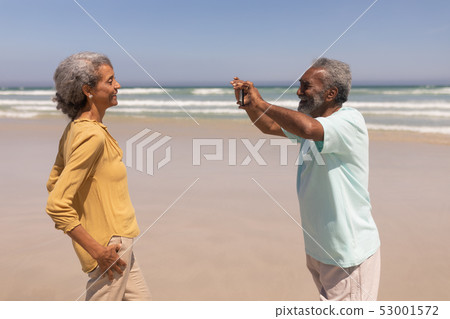Senior man clicking photo of woman with mobile phone on beach 53001572