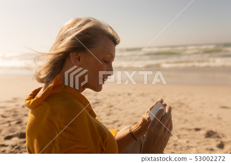 Senior woman reviewing through photos on mobile phone at beach 53002272
