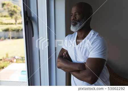 Senior man looking through window with arms crossed at home Senior man looking through window with arms crossed at home 53002292