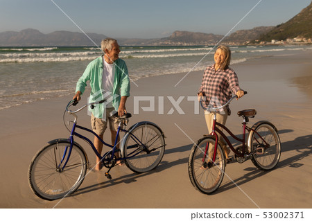 Senior couple standing with bicycle on the beach Senior couple standing with bicycle on the beach 53002371