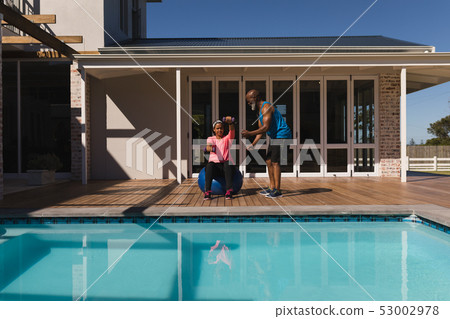 Male trainer training woman in balancing on exercise ball with dumbbells in the backyard Male trainer training woman in balancing on exercise ball with dumbbells in the backyard 53002978
