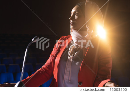 Businesswoman looking and thinking while standing on stage in auditorium Businesswoman looking and thinking while standing on stage in auditorium 53003300