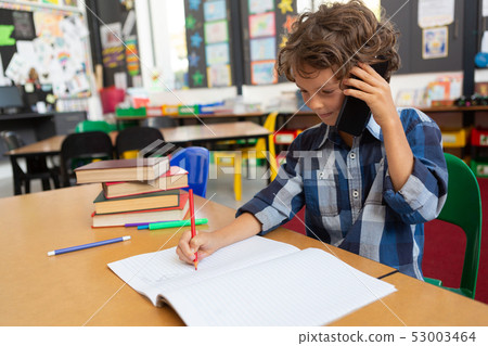 Schoolboy talking on mobile phone while writing in classroom Schoolboy talking on mobile phone while writing in classroom 53003464