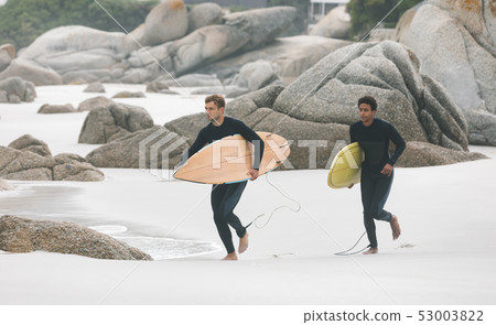 Male friends running on beach while holding surfboard 53003822