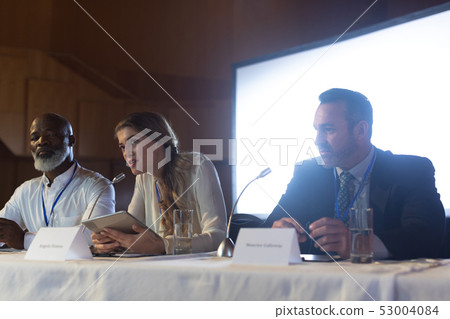 Businesswoman with executive speaking in speaker while sitting in the auditorium Businesswoman with executive speaking in speaker while sitting in the auditorium 53004084