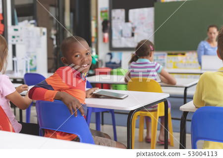 Happy schoolboy looking at the camera and holding digital tablet at desk in classroom Happy schoolboy looking at the camera and holding digital tablet at desk in classroom 53004113