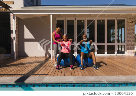 Female trainer training senior couple in performing exercise in the backyard Female trainer training senior couple in performing exercise in the backyard 53004130