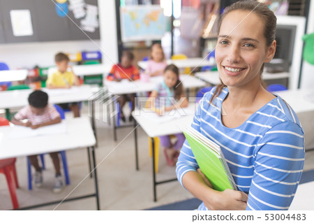 Happy schoolteacher standing in classroom with book Happy schoolteacher standing in classroom with book 53004483