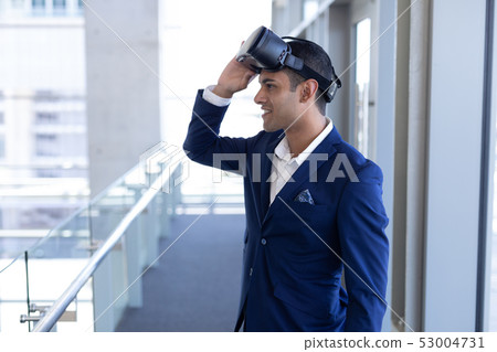 Mixed-race businessman using virtual reality headset standing in modern office 53004731