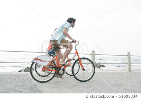 Caucasian couple riding bicycle on pavement near promenade at beach Caucasian couple riding bicycle on pavement near promenade at beach 53005334