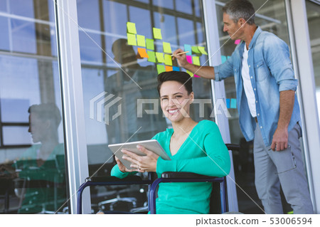 Disabled female executive  smiling while looking at camera while male executive writing on sticky no 53006594