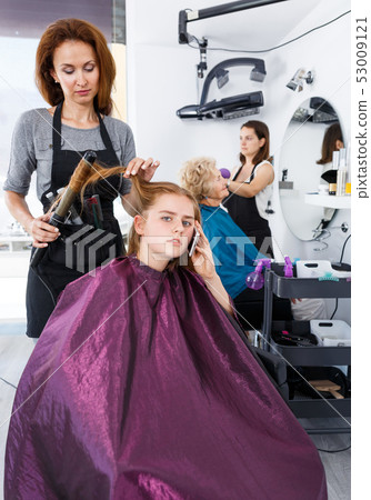 Stylist curling hair of teen girl with phone 53009121