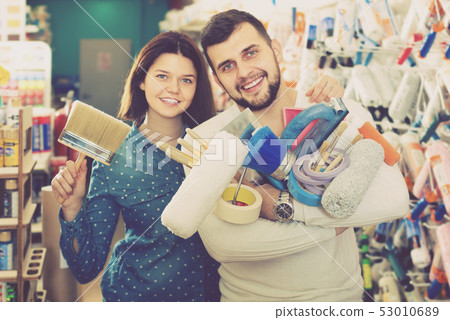 Young cheerful couple demonstrating tools for house renovating in paint store Young cheerful couple demonstrating tools for house renovating in paint store 53010689