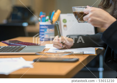 Closeup businesswoman hand writing and drinking coffee when working with with computer laptop and office supplies in modern office or meeting room, Business and worker concept 53011210