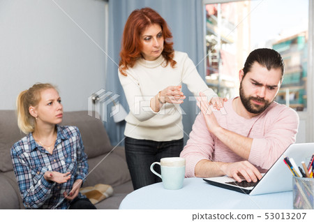 Upset young man working at laptop, young sister and mother Upset young man working at laptop, young sister and mother 53013207