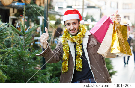 Man in Christmas hat with purchases delighted at Christmas fair, thumbs up Man in Christmas hat with purchases delighted at Christmas fair, thumbs up 53014202