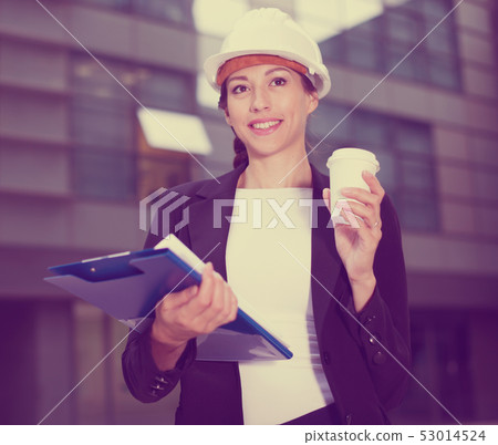 Female designer in suit and hat with folder is looking into documents with project and drinking coffee Female designer in suit and hat with folder is looking into documents with project and drinking coffee 53014524