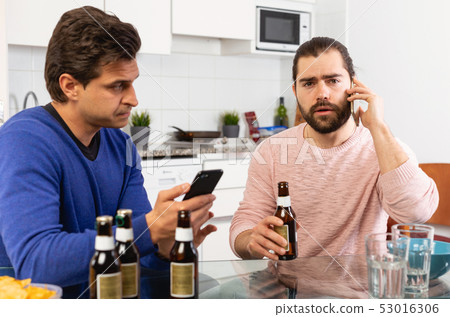 Two concerned men talking and laughing while enjoying beer at home, using phone Two concerned men talking and laughing while enjoying beer at home, using phone 53016306