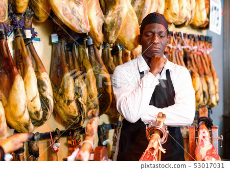 Cheerful African American seller showing thumbs up while offering delectable spanish jamon in butcher shop Cheerful African American seller showing thumbs up while offering delectable spanish jamon in butcher shop 53017031