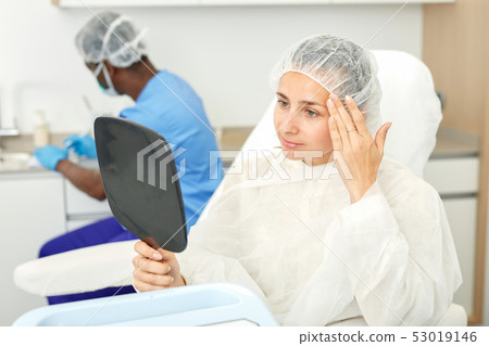 Young woman sitting in armchair and looking to mirror before mesotherapy Young woman sitting in armchair and looking to mirror before mesotherapy 53019146