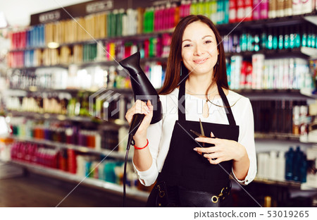 Female hairdresser in apron holding blow dryer and hair cutters in shop Female hairdresser in apron holding blow dryer and hair cutters in shop 53019265