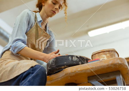 Attentive curly girl in dirty apron working on special pottery wheel Attentive curly girl in dirty apron working on special pottery wheel 53026738