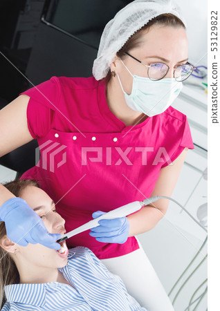 A professional dentist woman in glasses and overalls examines the oral cavity of a young girl in the 53129822