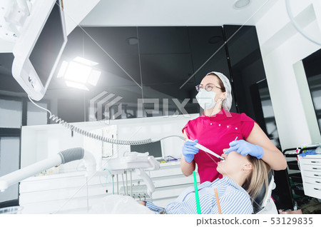 A professional dentist woman in glasses and overalls examines the oral cavity of a young girl in the A professional dentist woman in glasses and overalls examines the oral cavity of a young girl in the 53129835