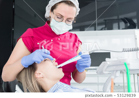 A professional dentist woman in glasses and overalls examines the oral cavity of a young girl in the A professional dentist woman in glasses and overalls examines the oral cavity of a young girl in the 53129839