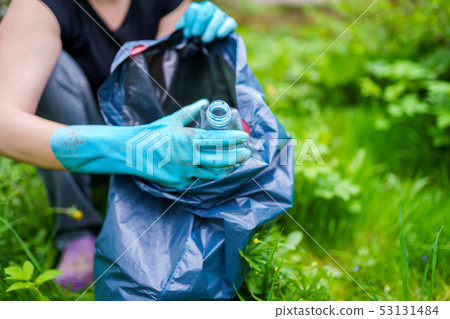 Image of woman in rubber gloves picking up dirty plastic bottle in bag on green lawn 53131484