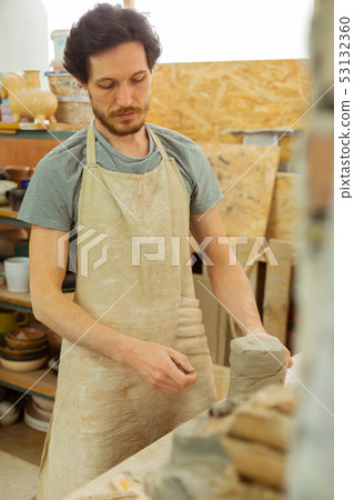 Focused dark-haired man in special apron measuring needed amount of clay Focused dark-haired man in special apron measuring needed amount of clay 53132360